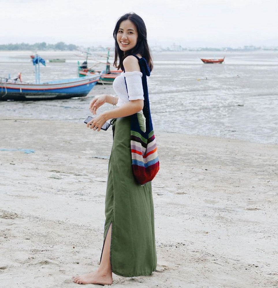 Woman in green Thai wrap pants skirt and white top standing on a beach with ocean waves and a boats in the background