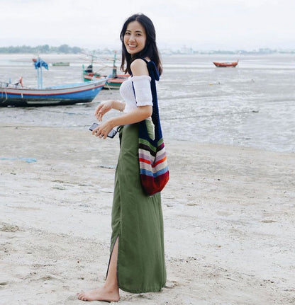 Woman in green Thai wrap pants skirt and white top standing on a beach with ocean waves and a boats in the background