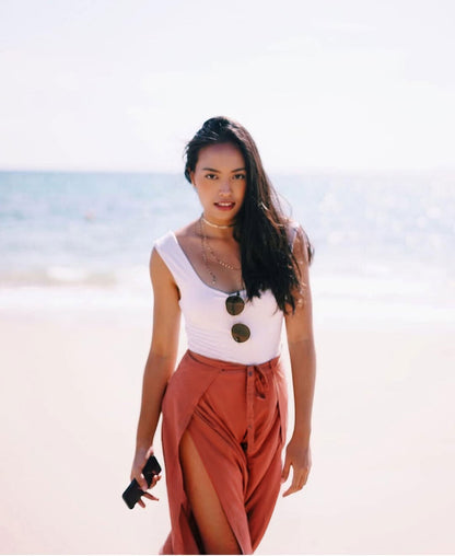 Woman standing on a beach wearing a white top and orange thai wrap pants skirt.