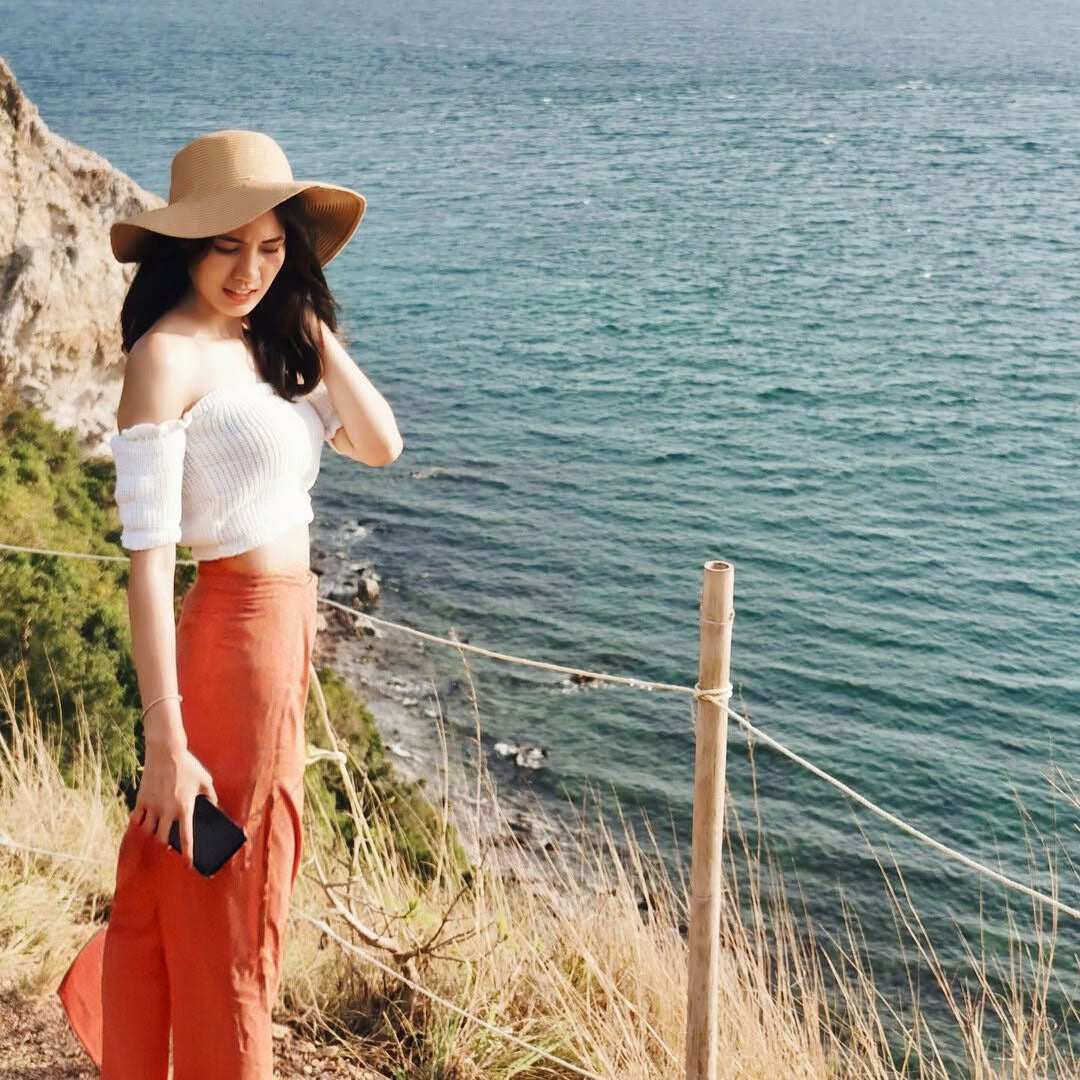 Woman in a white top and orange thai wrap pants skirt standing on a cliff overlooking the ocean.