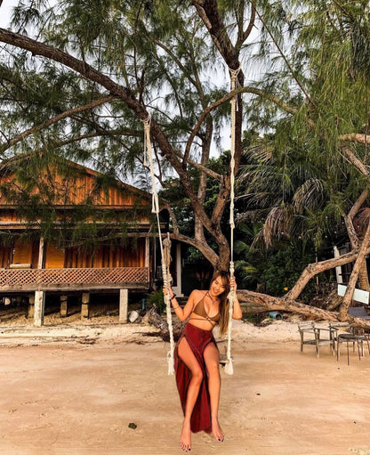 Woman on a swing in a tropical setting with wooden hut and trees wearing a maroon Thai Wrap Pants Skirt 