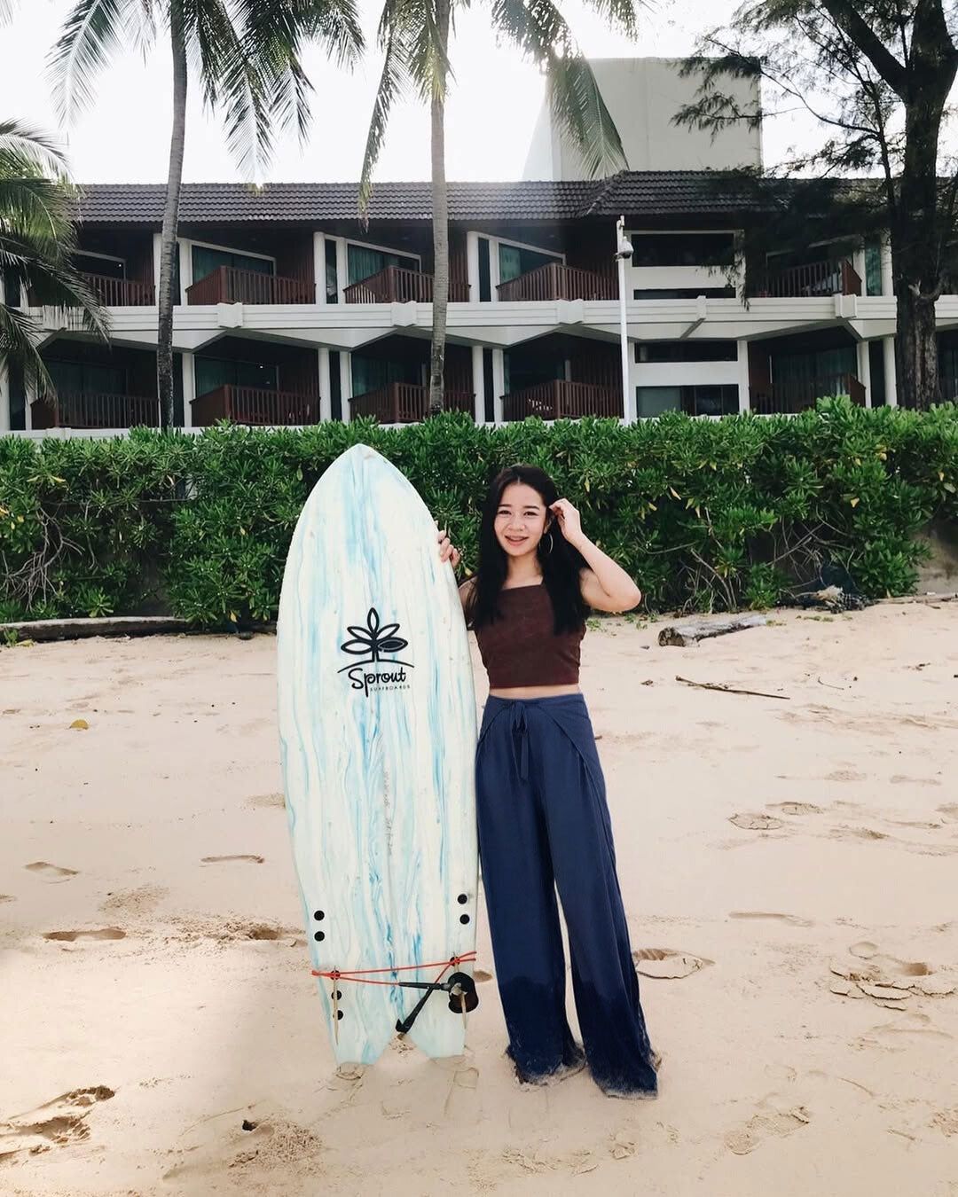 Woman in blue Thai wrap pants skirt and brown top holding a surfboard on a beach with a building and palm trees in the background
