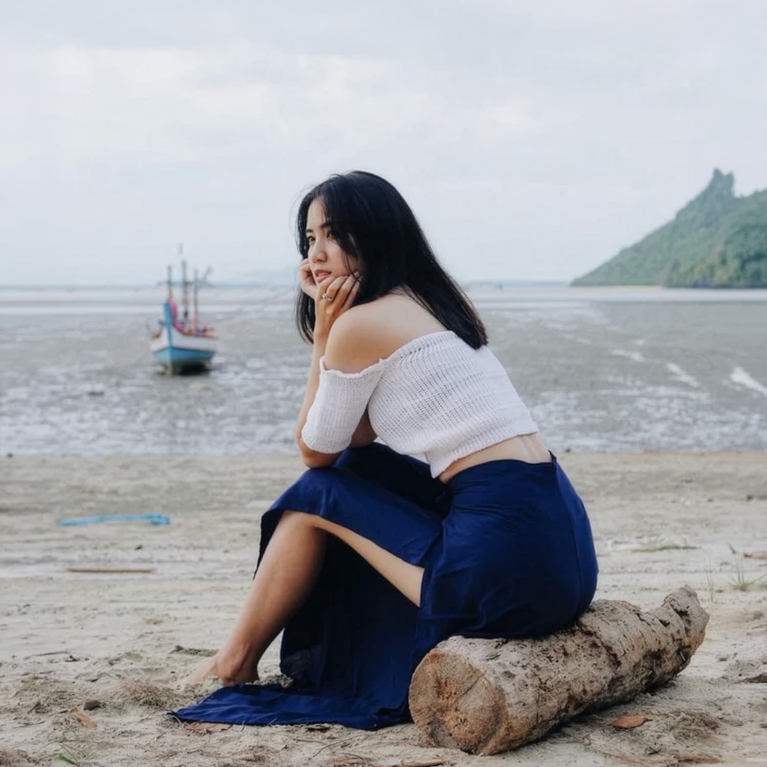 Woman in blue Thai wrap pants skirt and white top sitting on a log by the water with a boat in the background