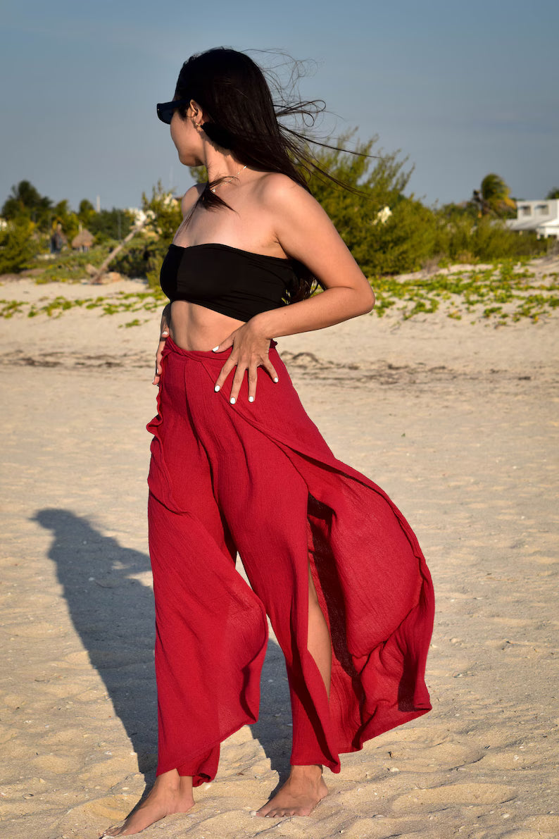 Woman in black top and red thai wrap pants skirt standing on a sandy beach with greenery in the background