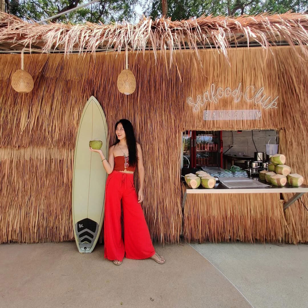 Woman in red Thai wrap pants skirt and red top holding a coconut next to a surfboard in front of a thatched roof building 