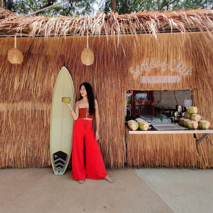 Woman in red Thai wrap pants skirt and red top holding a coconut next to a surfboard in front of a thatched roof building 
