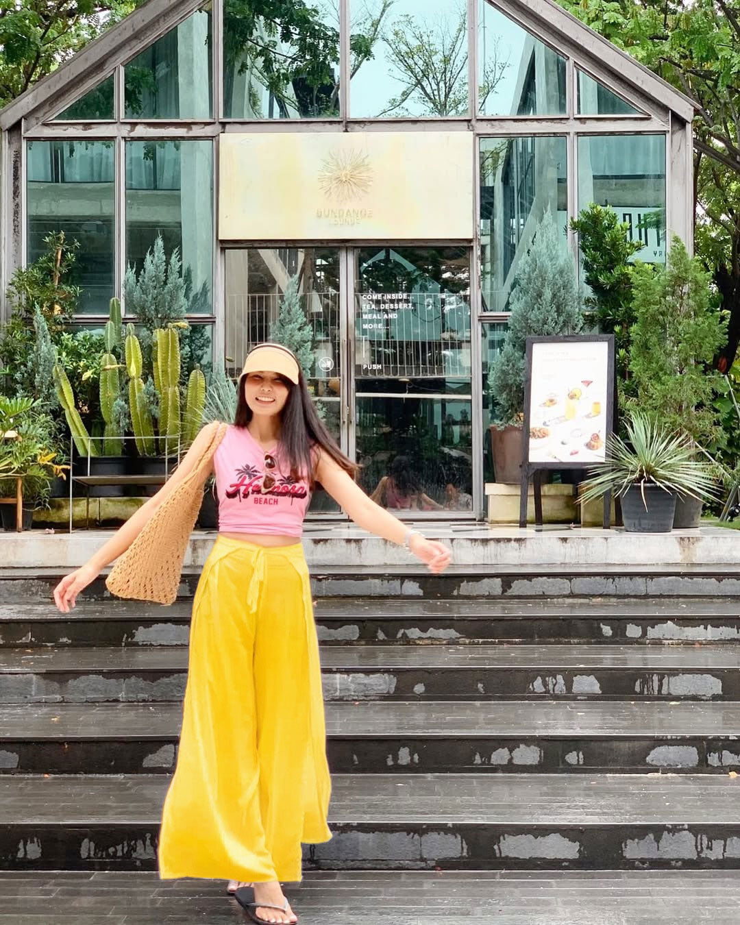 Woman in pink top and yellow thai wrap pants skirt standing on steps with plants and a building in the background