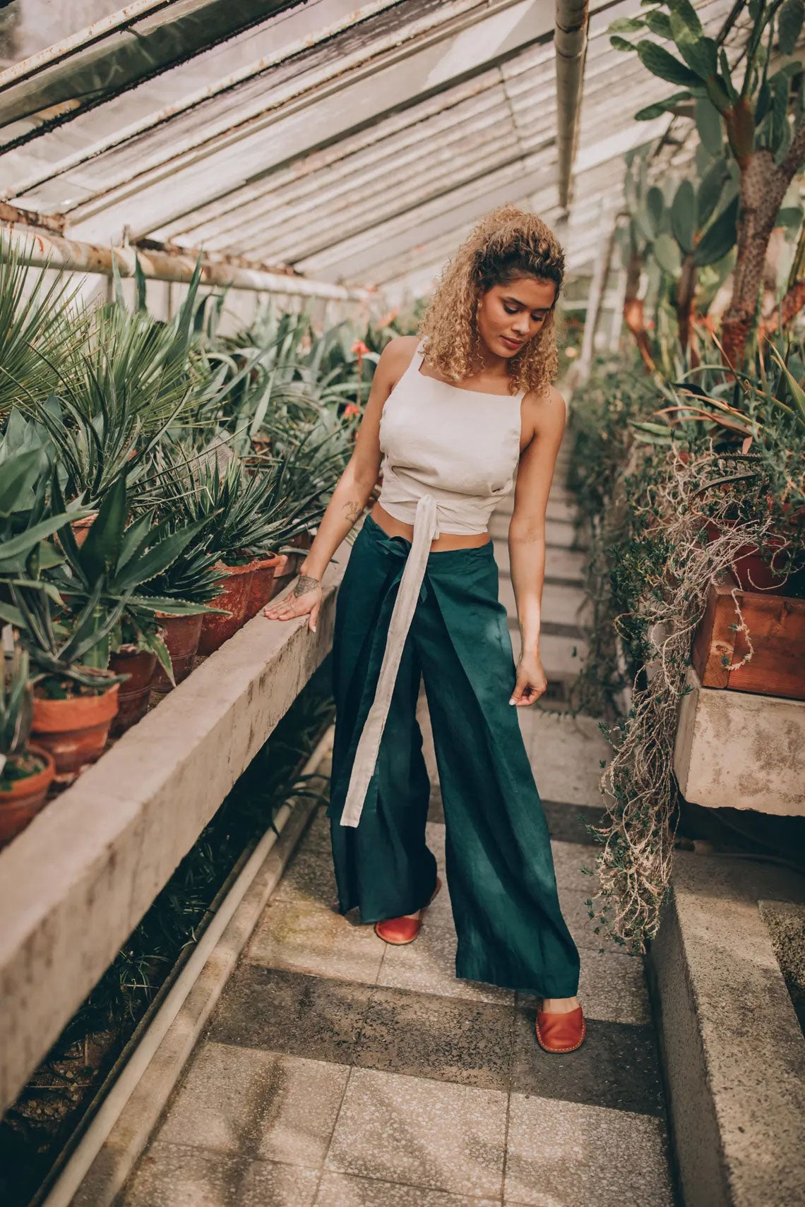Woman in a white top and a turquoise Thai Wrap Pants Skirt walking through a greenhouse.