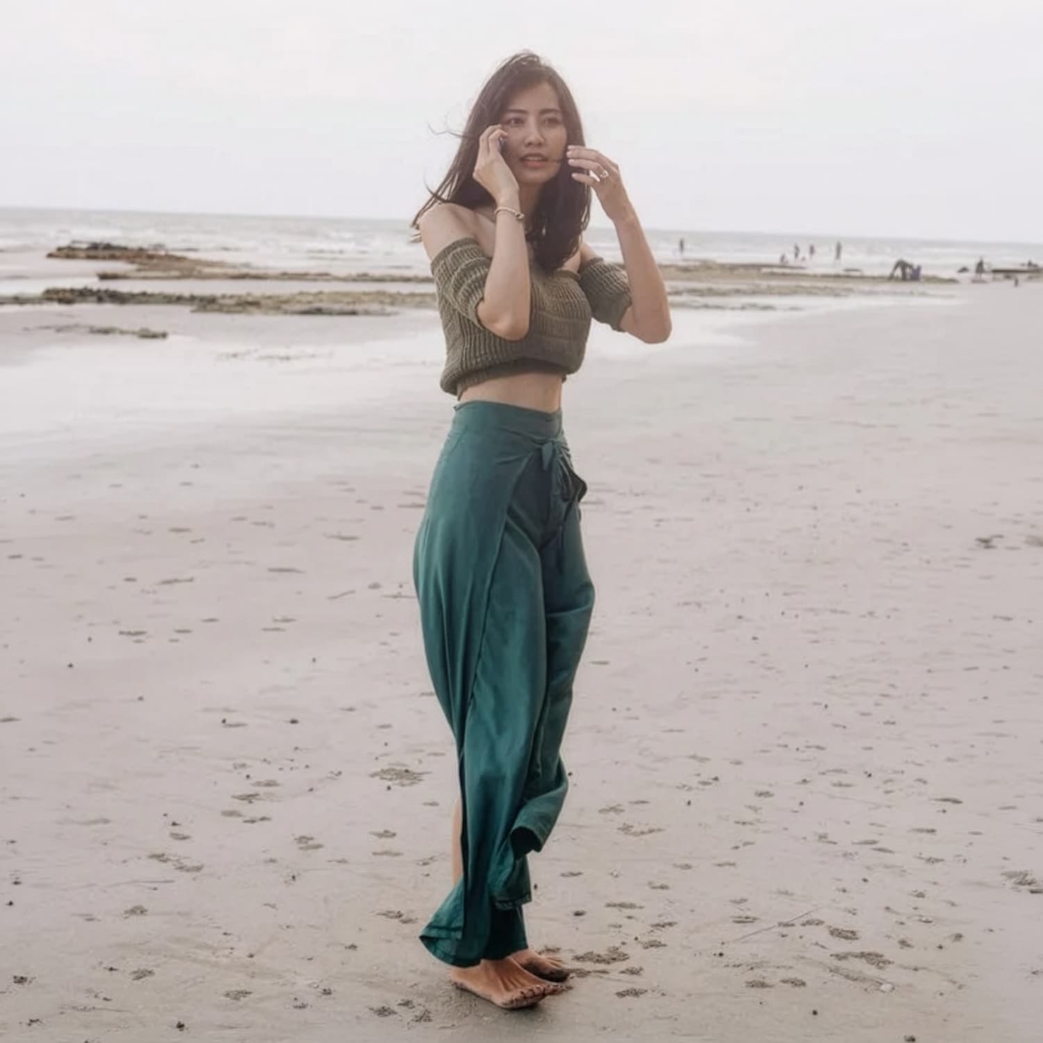 Woman in turquoise thai wrap pants skirt and brown top standing on a beach with rocks in the background