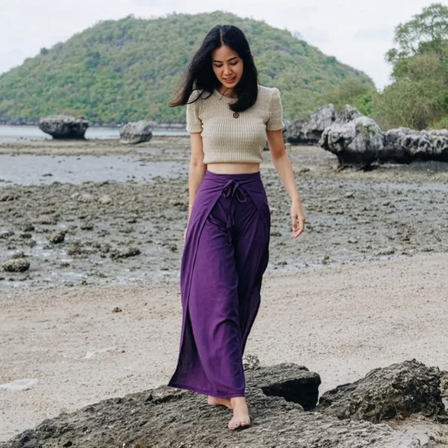 Woman in purple thai wrap pants skirt and beige top standing on a beach with rocks in the background