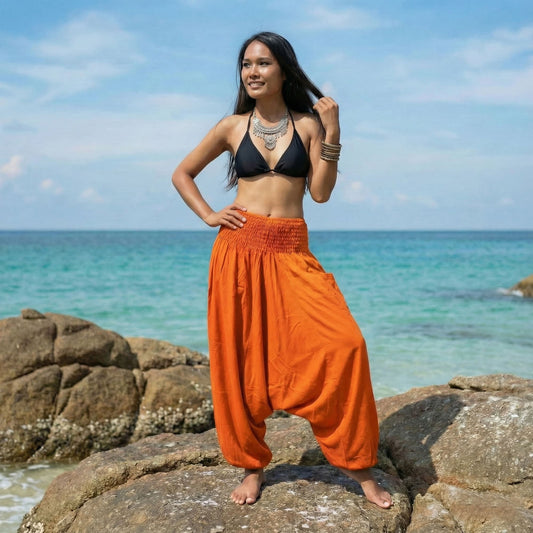 Woman in black bikini top and orange thai harem pants jumpsuit standing on rocks by the ocean