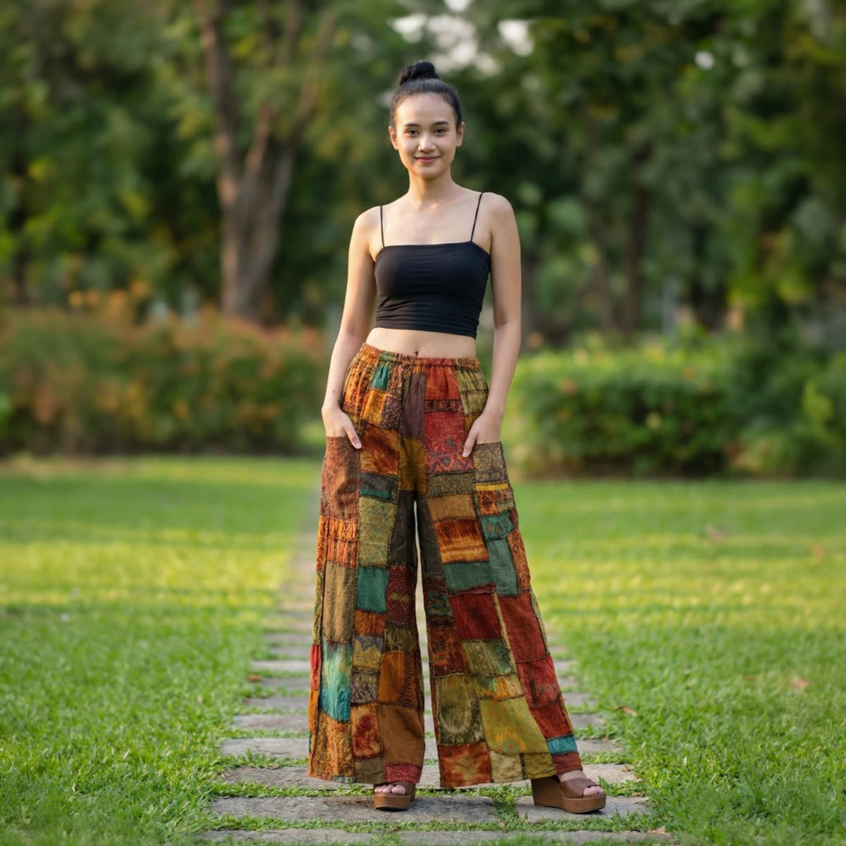 Woman in orange patchwork wide-leg boho palazzo pants standing in a sunny park, front view.