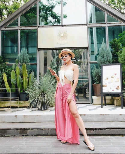 Woman in a white top and pink thai wrap pants skirt standing outdoors with plants and a building in the background