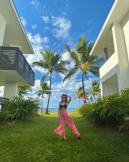 Woman in pink thai wrap pants skirts and black top standing in a tropical garden with palm trees and modern buildings.