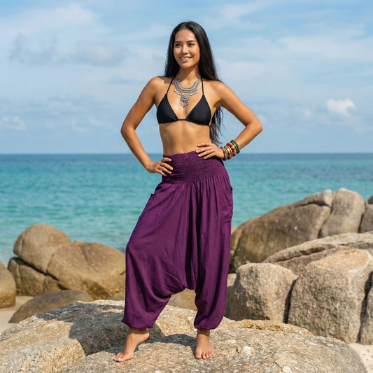 Woman in black bikini top and purple jumpsuit thai pants standing on rocks with ocean and sky in the background