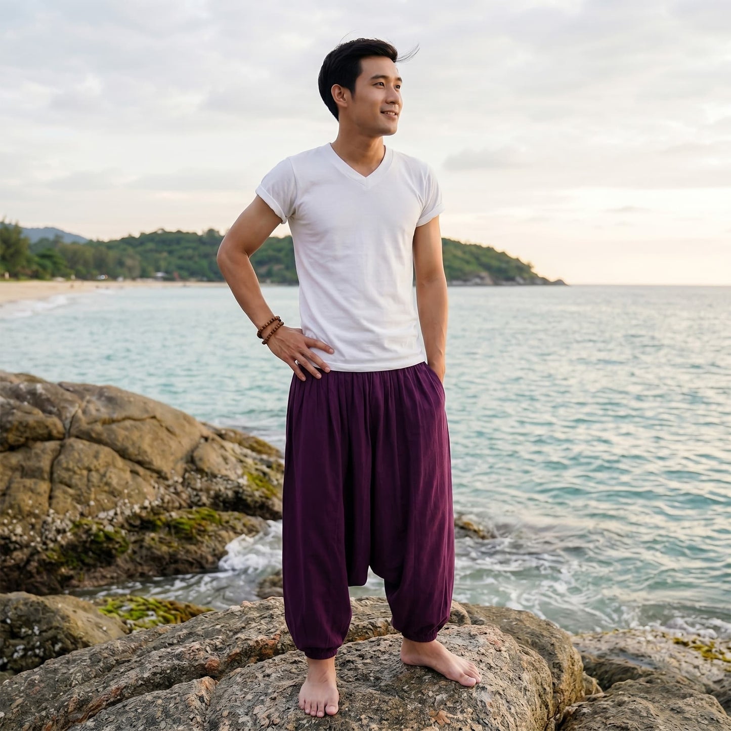 Male model standing on rocks by the ocean wearing deep purple Men's drop crotch harem pants.