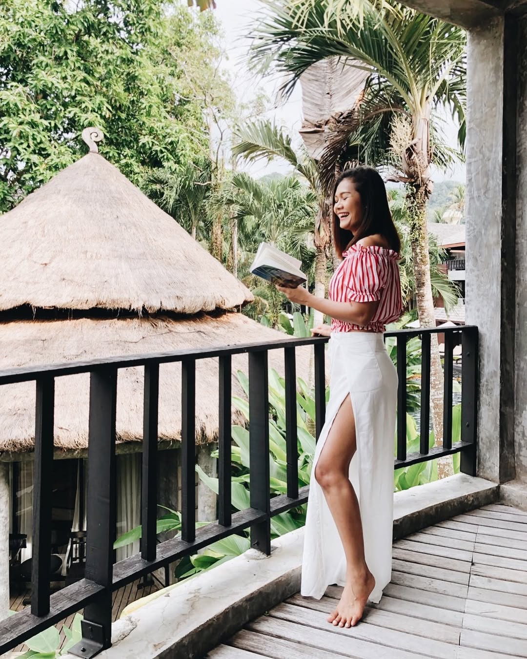 Woman n white thai wrap pants skirt and red top standing on a balcony with tropical greenery in the background