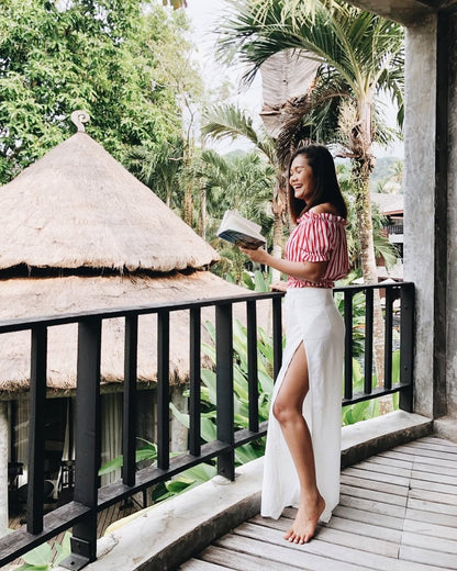 Woman n white thai wrap pants skirt and red top standing on a balcony with tropical greenery in the background