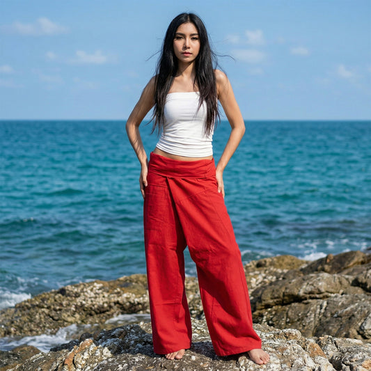 Female model standing confidently on rocks by the ocean, wearing versatile Crimson Sunset women's Thai fisherman pants.
