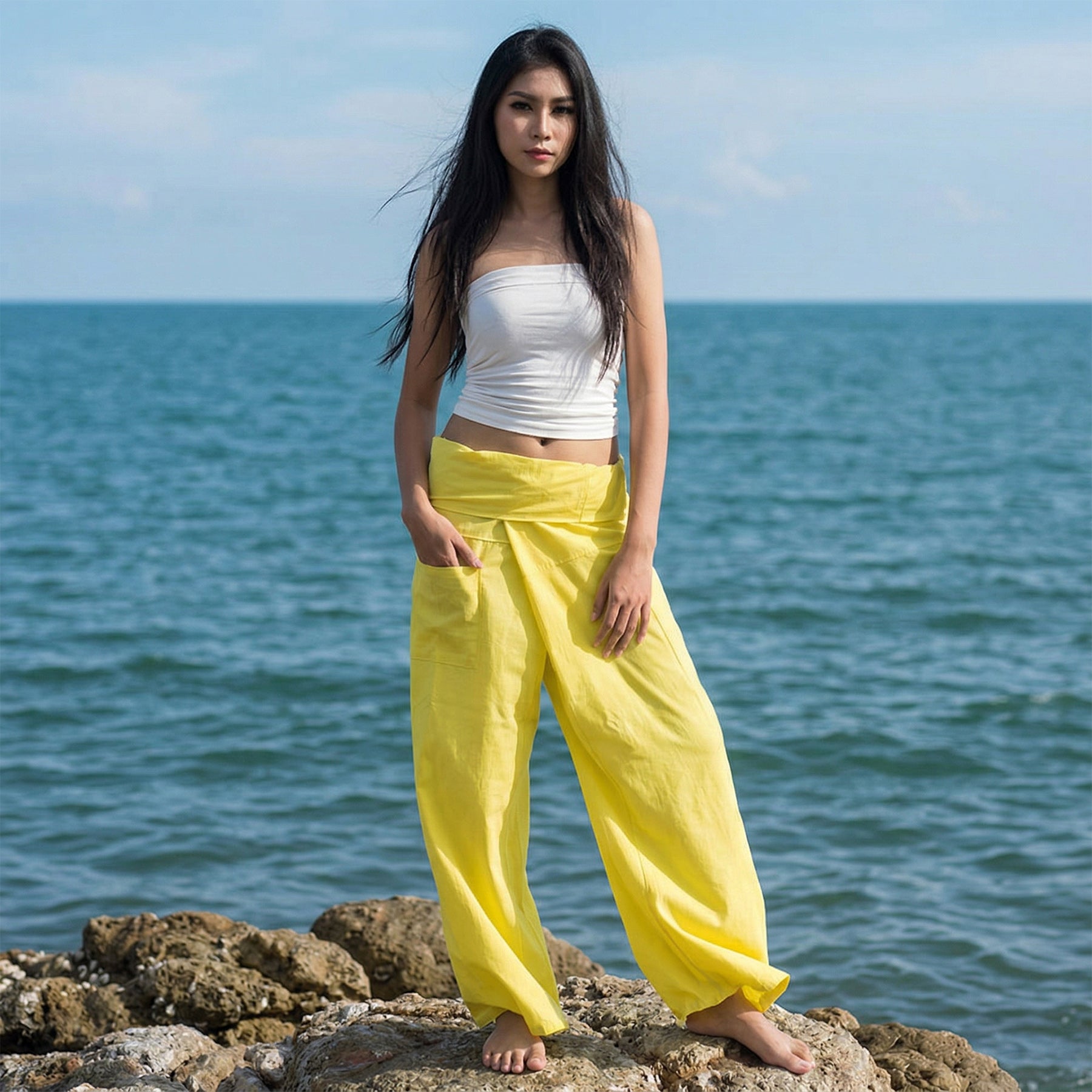Female model standing confidently on rocks by the ocean, wearing versatile Golden Sunshine women's Thai fisherman pants.