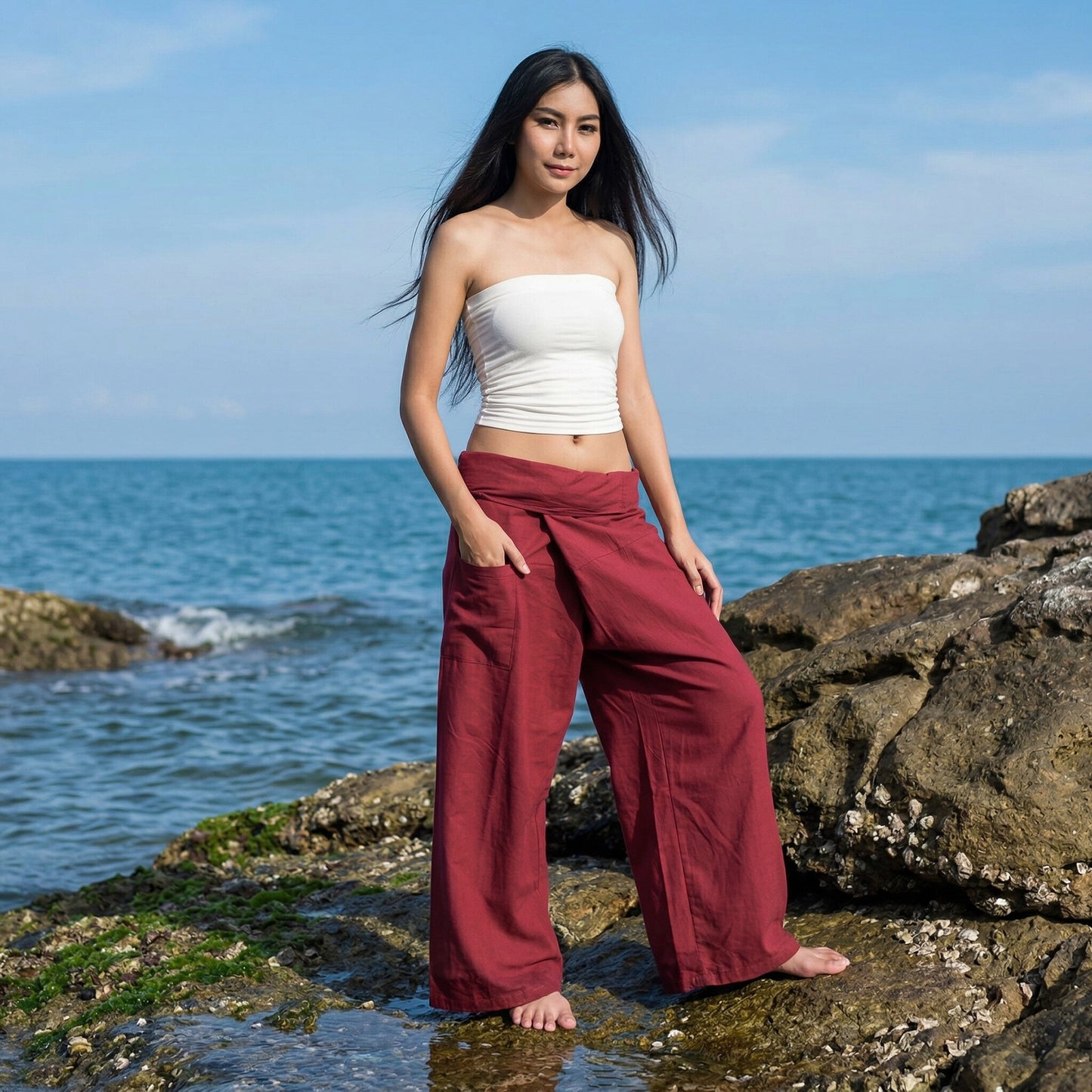 Female model standing confidently on rocks by the ocean, wearing versatile Mystic Maroon women's Thai fisherman pants.