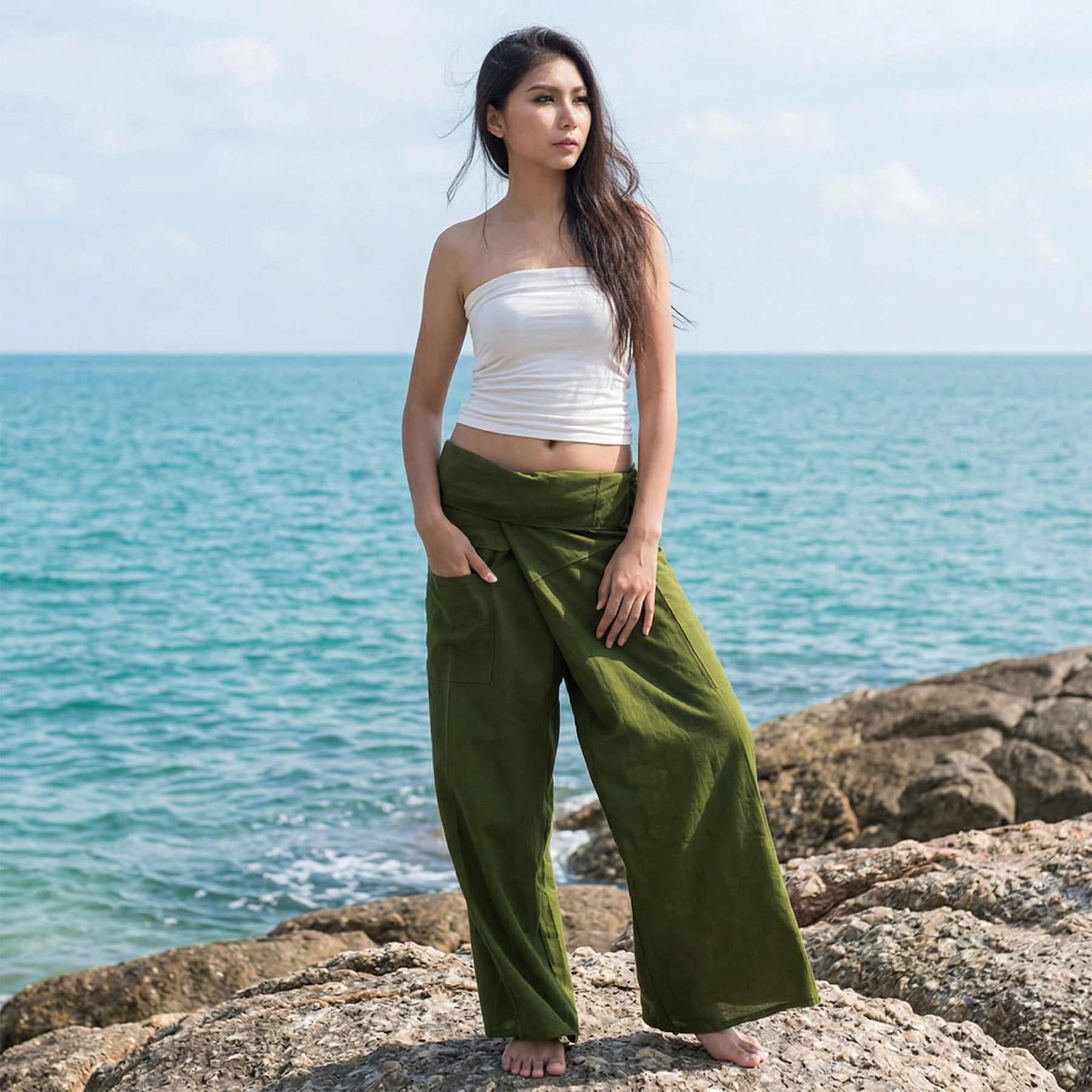 Female model standing confidently on rocks by the ocean, wearing versatile Olive Grove women's Thai fisherman pants.