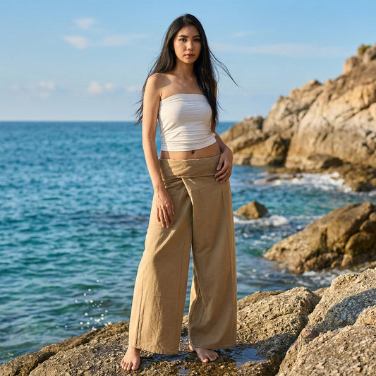 Female model standing confidently on rocks by the ocean, wearing versatile Sandy Beige women's Thai fisherman pants.