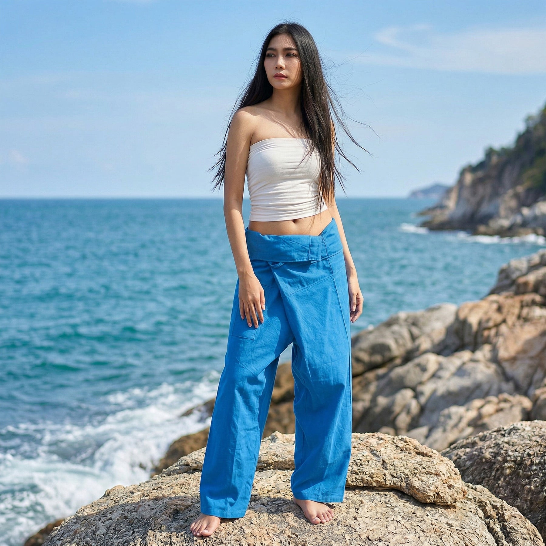 Female model standing confidently on rocks by the ocean, wearing versatile Sky Blue Horizon women's Thai fisherman pants.