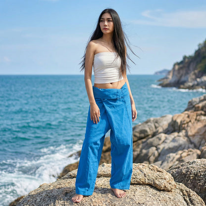 Female model standing confidently on rocks by the ocean, wearing versatile Sky Blue Horizon women's Thai fisherman pants.