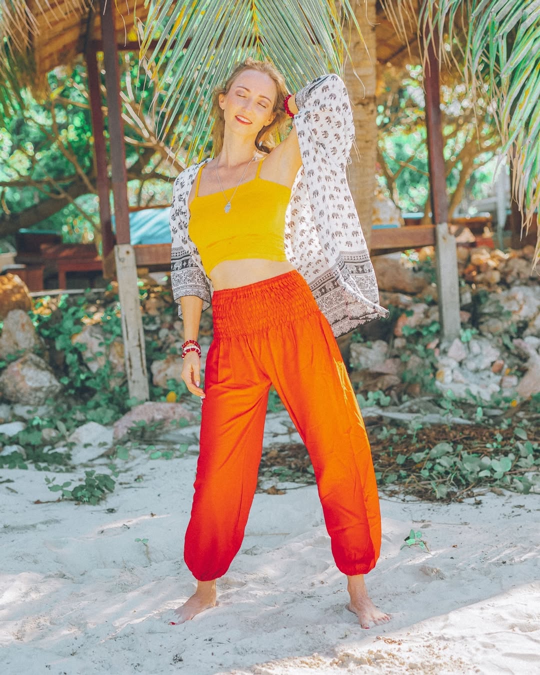 Woman in yellow top and red my thai harem pants standing on a sandy beach with tropical plants in the background