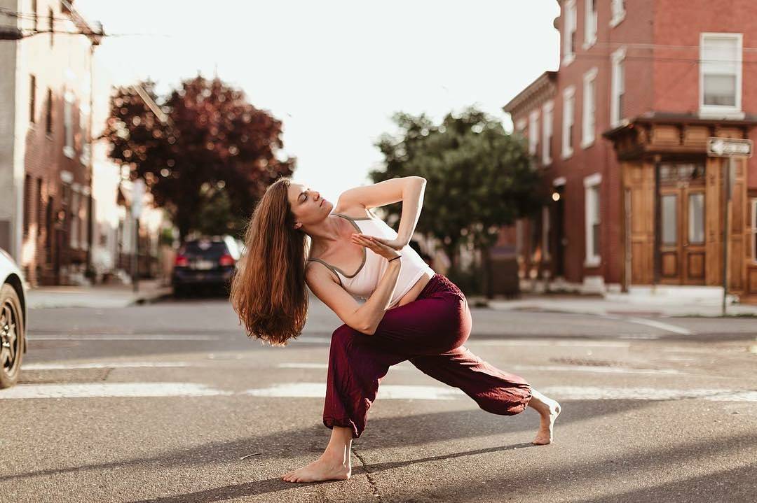 Woman practicing yoga my thai harem pants on a city street with buildings and trees in the background