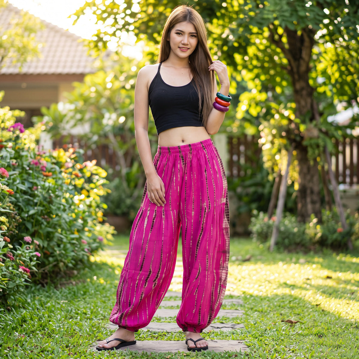 Female model wearing bright pink Lotus Bloom Tie Dye Harem Pants while standing among lush plants in a backyard garden.