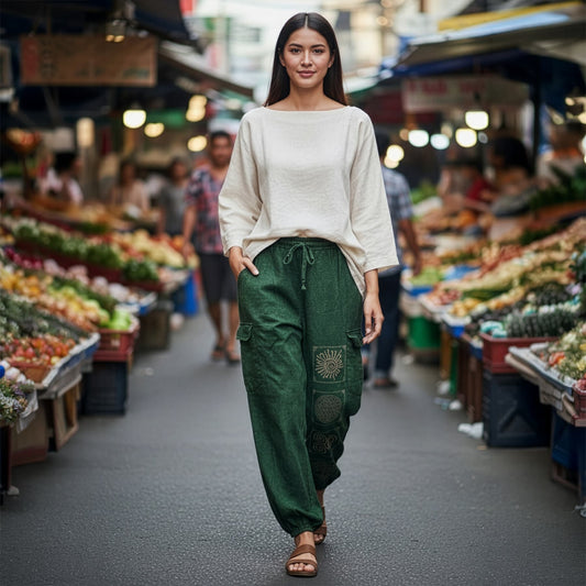 Female model wearing green boho tribal mandala harem pants at a busy Thai market, front view.
