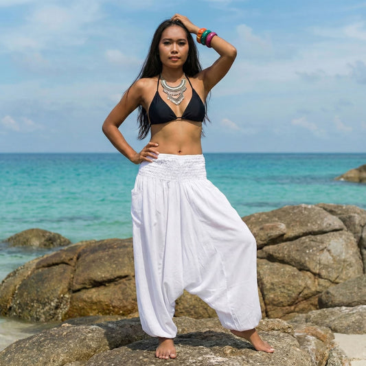 Woman in black bikini top and white thai harem pants jumpsuit standing on rocks by the ocean
