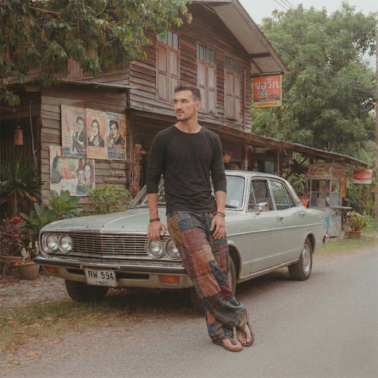 Male model wearing black boho patchwork pants leaning on a classic car in front of an old Thai grocery store.