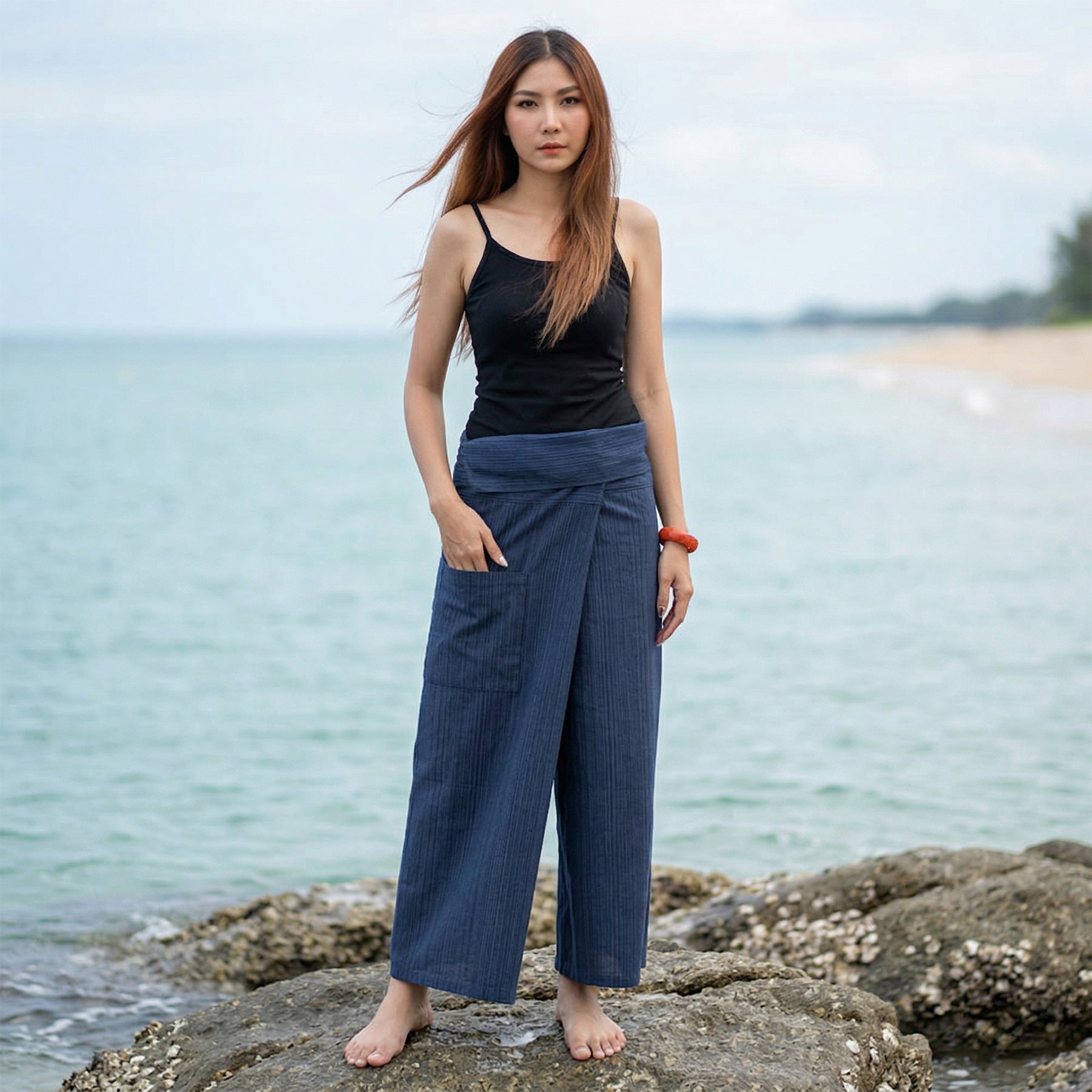 Female model standing confidently on rocks by the ocean, wearing versatile Blue Lagoon women's pinstripe Thai fisherman pants.