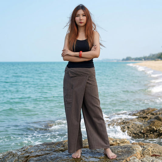 Female model standing confidently on rocks by the ocean, wearing versatile Earthy Brown women's pinstripe Thai fisherman pants.