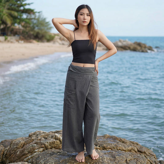 Female model standing confidently on rocks by the ocean, wearing versatile Elephant Gray women's pinstripe Thai fisherman pants.
