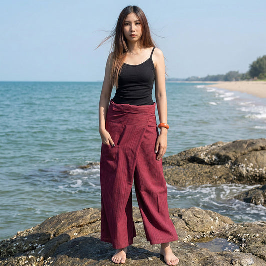 Female model standing confidently on rocks by the ocean, wearing versatile Sunset Maroon women's pinstripe Thai fisherman pants.