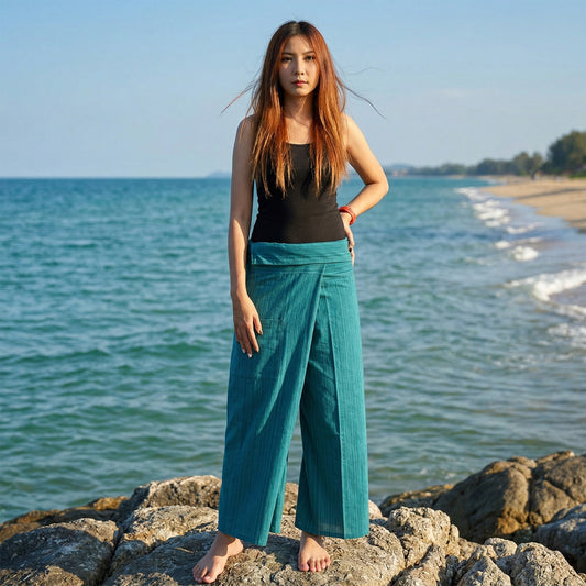 Female model standing confidently on rocks by the ocean, wearing versatile Turquoise Bay women's pinstripe Thai fisherman pants.